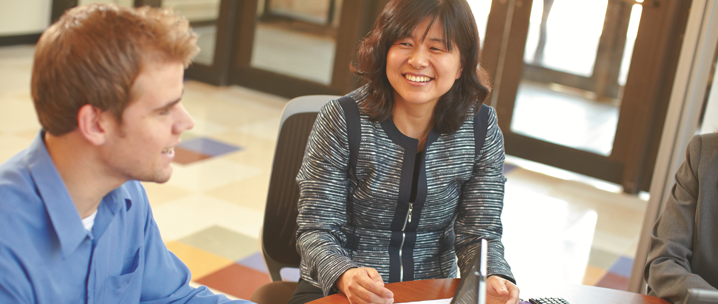 Picture of female, Asian nontraditional student smiling, sitting next to Caucasian male student in what appears to be a classroom