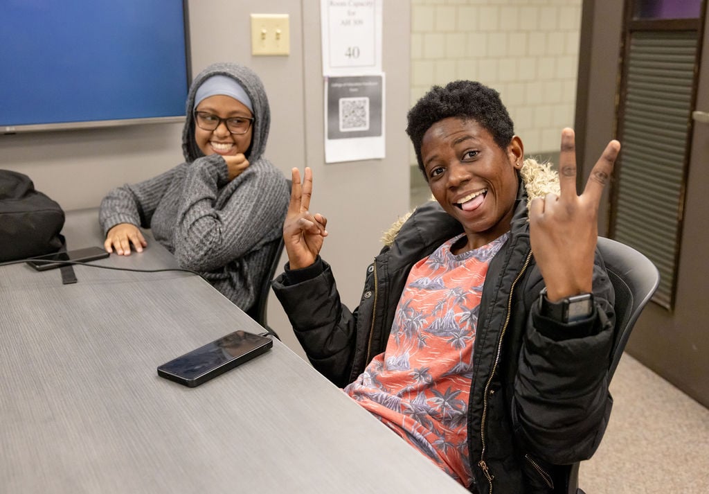 Two students sitting at a desk in an MSU classroom. One student looks at the other and smiles. The other student is giving two peace signs and making a funny face at the camera.