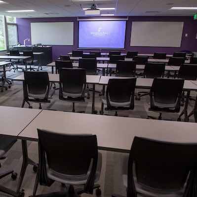 Classroom with tables and chairs and a display screen up front with whiteboards on both sides.