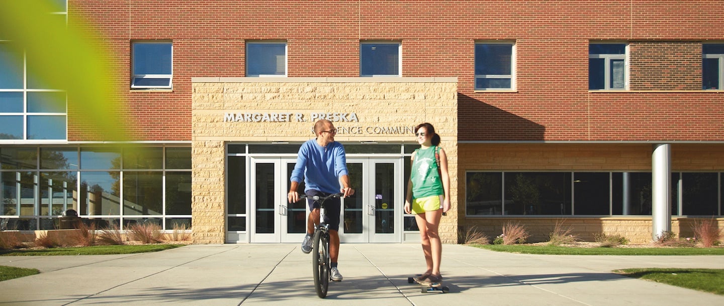 A boy bicycling with a girl skating in front of the Preska Community building