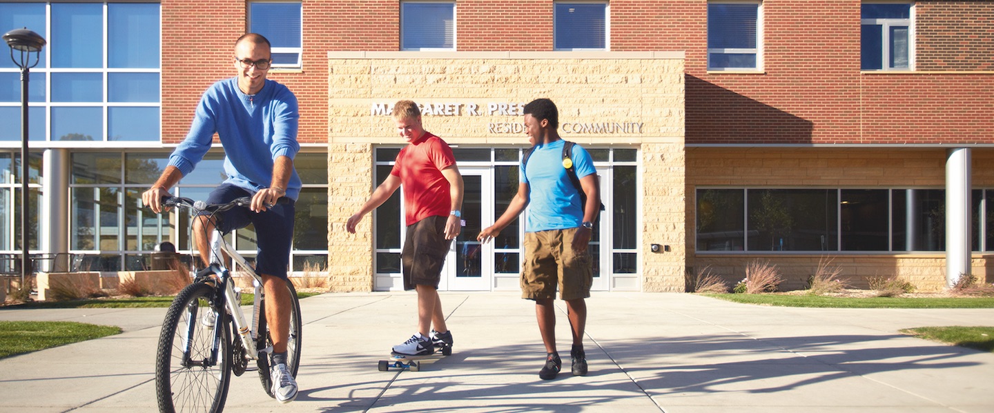 3 students, 1 guy is bicycling, 1 guy is skating and 1 guy is wallking in front of the Preska Residance Hall building