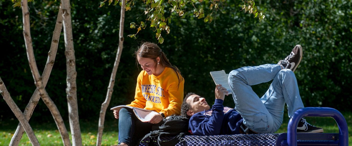 A student sitting and another student lying down on a bench outside studying on a sunny day
