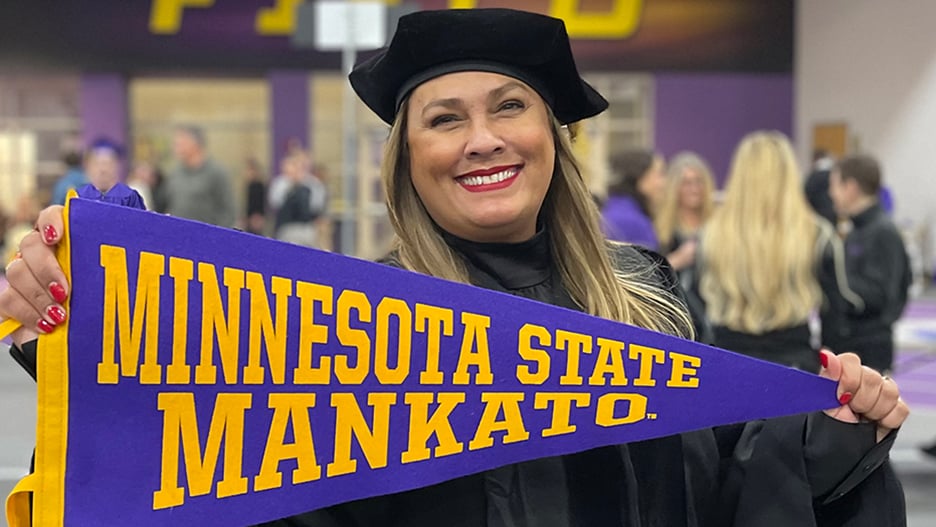 Isabel Rodriguez-Mendoza, the College of Education’s first Latina doctoral graduate, posing in her cap and gown holding a Minnesota State Mankato banner after the commencement ceremony