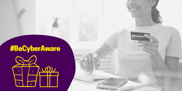 Woman smiling and using credit card to purchase online. Gold gift icons and text that says: "#BeCyberAware"