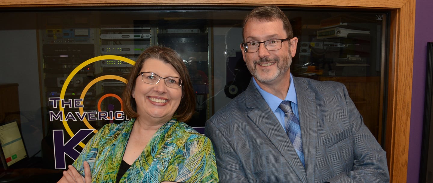 Karen Wright and Dwayne Megaw smiling and posing in front of the KMSU Radio station studio window 