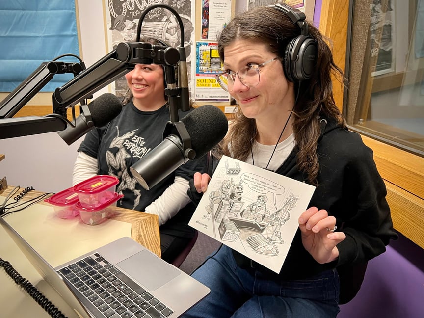 Amber Rahe and Kat Baumann, artists that helped during the pledge drive. Kat is holding her illustration of Shyboy Tim and Shelley being held hostage by DJ Transmitter