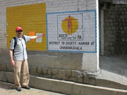 Zorba Paster standing by an older building pointing at the wall with a painted message that says "DOTS sure cure for TB. District TB Society Kangra at Dharamshala"