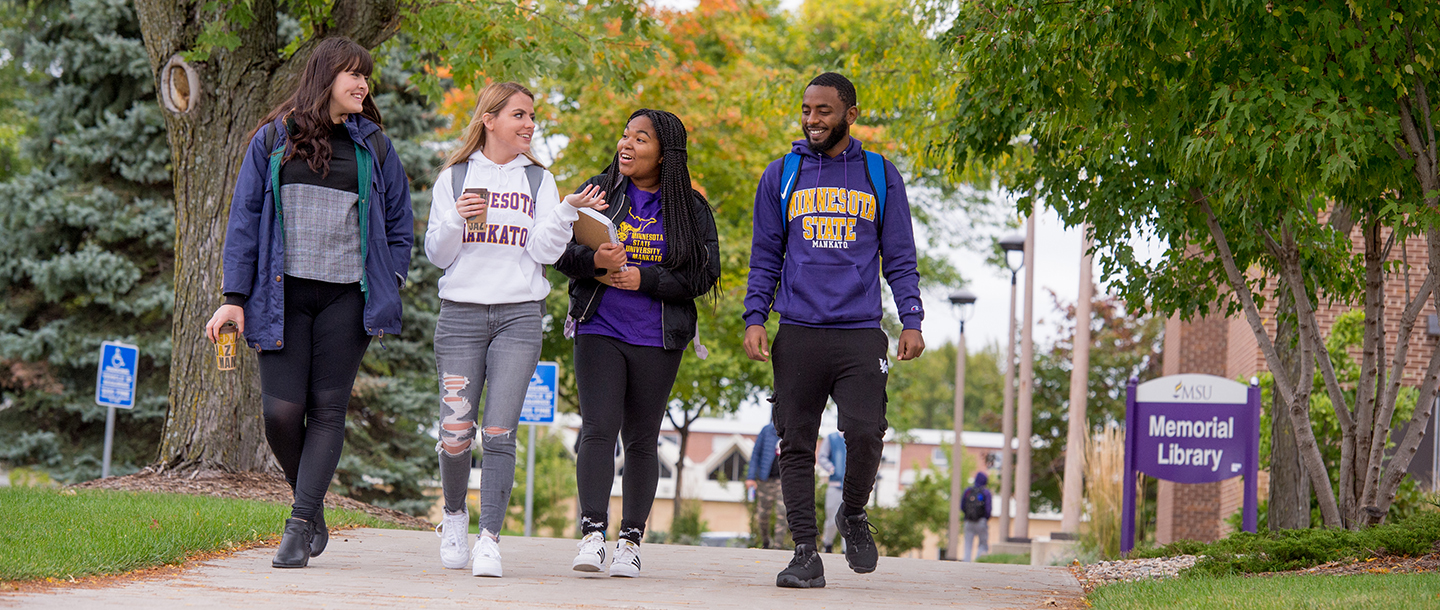 Four students walking on a sidewalk in front of the Memorial Library.