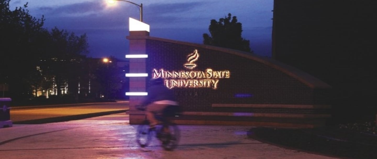 A person riding a bicycle in front of the Minnesota State University logo at the entrance of campus at night