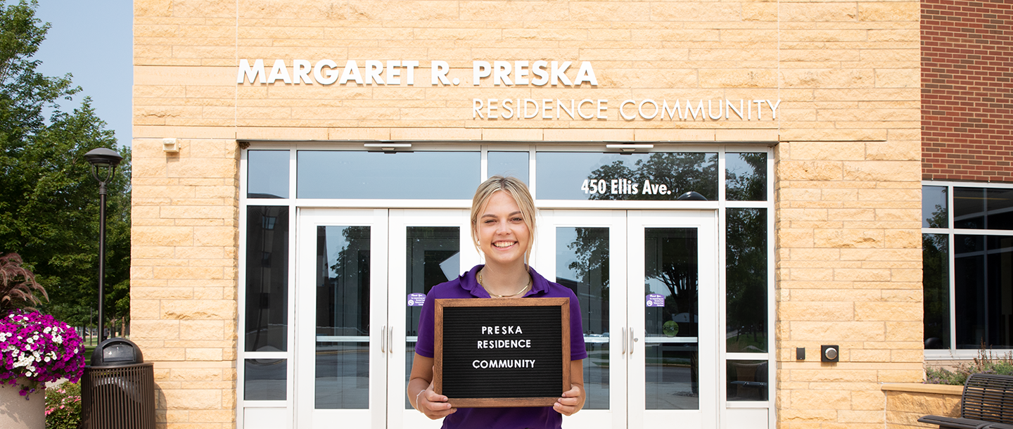 A student, standing in front of Preska residence community, holding a sign that says Preska Residence Community