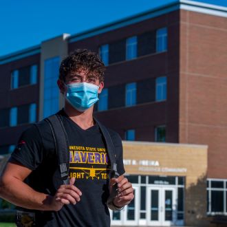 Male student with a mask on standing outside in front of the Margaret R. Preska Residence Community building